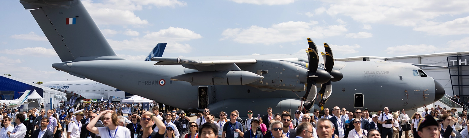 Visiteurs professionnels découvrant les démonstrations aériennes avec un avion de transport militaire en fond, exposé au Salon International de l’Aéronautique et de l’Espace à Paris-Le Bourget.